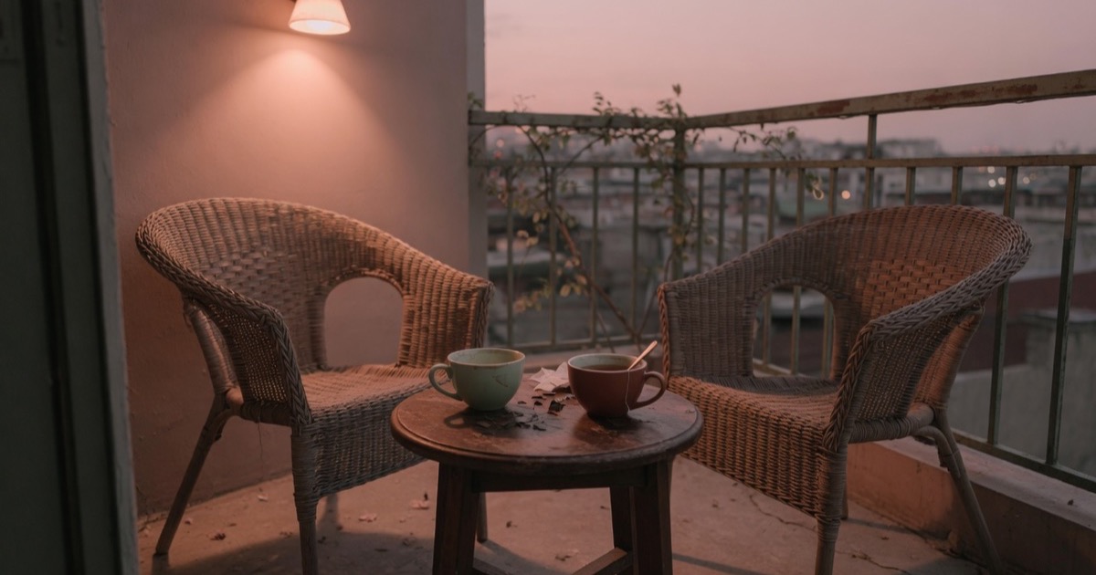 Due poltrone vuote una di fronte all'altra su un balcone, con un tavolino e due tazze di tè, nella luce calda del tramonto.