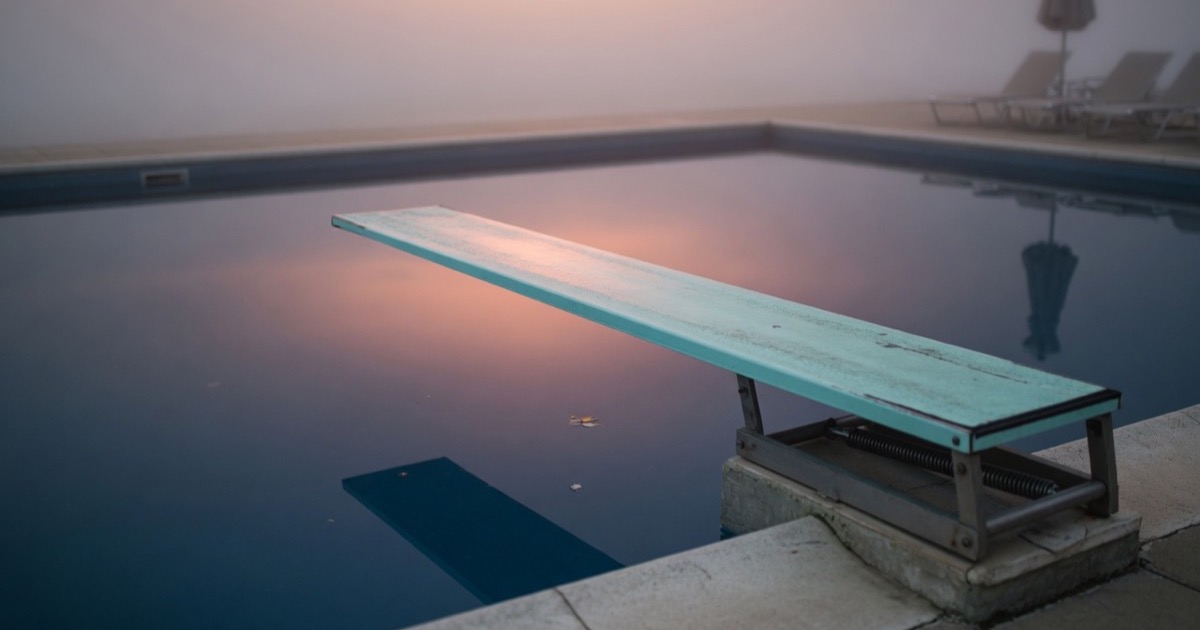An empty diving board stretching out over still pool water in early morning light.