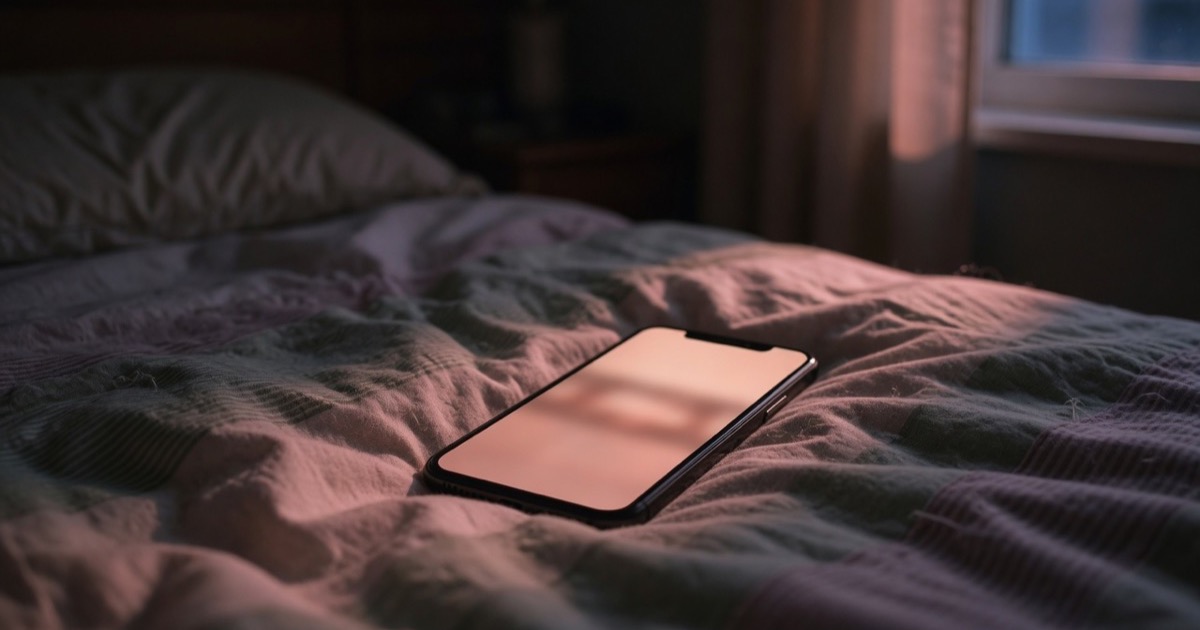Ein Smartphone mit hellem Display liegt auf einem unaufgeräumten Bett in einem dunklen Zimmer, spät in der Nacht.